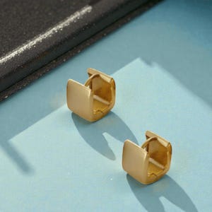 May include: Two gold-colored square hoop earrings. The earrings have a matte finish and a simple, modern design. They are displayed on a light blue surface, with a dark blue and black object in the background.