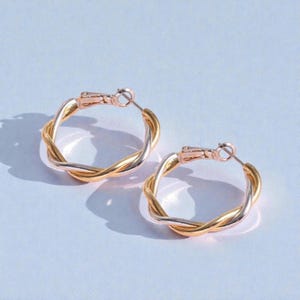 Puede incluir: Un par de pendientes de aro trenzados en oro y plata. Los pendientes tienen un diseño trenzado con una combinación de colores oro y plata. Los aros se aseguran con un cierre de bisagra. Los pendientes se muestran sobre una superficie azul claro.