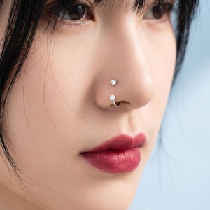 May include: A silver-toned nose piercing set consisting of two pieces: a small stud with a clear round gemstone and a curved hoop featuring a matching clear round gemstone at the end, both worn on the side of the nostril for a layered appearance.