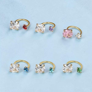 Puede incluir: Seis pendientes dorados de aro abierto. Cada uno tiene una piedra grande, transparente y de corte cuadrado y una piedra más pequeña de color. Las piedras de color son rojas, moradas, rosas, azules, turquesas y verdes. Sobre fondo azul claro.