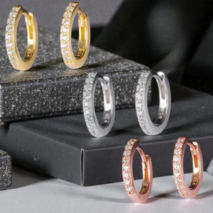 May include: An assortment of hoop earrings in gold, silver, and rose gold. Each pair is encrusted with small, sparkling stones. The earrings are displayed on a dark, textured surface, showcasing their different metallic finishes.