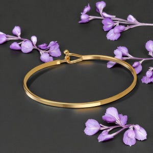 Puede incluir: Una pulsera dorada con cierre de gancho, sobre un fondo oscuro con flores moradas. La pulsera tiene un diseño sencillo y elegante. Las flores añaden un toque de color y contraste a la joya.