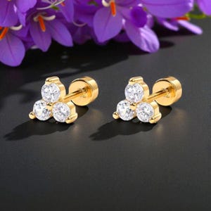 May include: A pair of gold-colored stud earrings, each featuring three clear, round cubic zirconia stones. The earrings are displayed on a black surface, with purple flowers in the background.