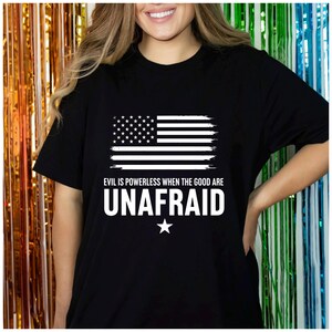 Może przedstawiać: Czarny t-shirt z białym, postarzanym wzorem amerykańskiej flagi. Koszulka zawiera napis "EVIL IS POWERLESS WHEN THE GOOD ARE UNAFRAID" w kolorze białym, z białą gwiazdą poniżej. Klasyczny okrągły dekolt.