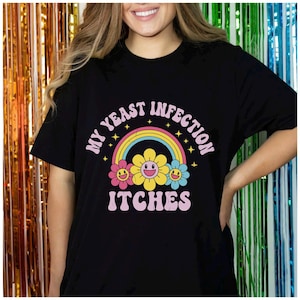以下が含まれることがあります： 黒いTシャツ。ピンクと白で「MY YEAST INFECTION ITCHES」の文字。デザインには、虹と黄色、ピンク、青の笑顔の花のグラフィックが含まれています。半袖のシャツです。
