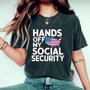May include: A dark gray t-shirt with the text "Hands Off My Social Security" in white letters. An American flag is displayed in the center of the shirt.