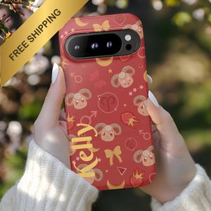 May include: Red phone case with a repeating pattern of Aries ram illustrations, stars, moons, and hearts. The name "Kelly" is printed in yellow script. The case is held in front of a blurred green background.