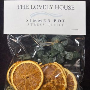 Puede incluir: Una bolsa de plástico transparente que contiene rodajas de naranja secas, hojas verdes y pequeñas hierbas verdes. La bolsa está etiquetada como "THE LOVELY HOUSE SIMMER POT STRESS RELIEF".