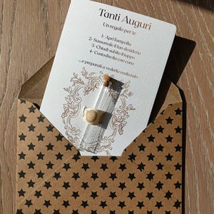 Può includere: Un piccolo flacone di vetro con tappo in sughero ed etichetta decorativa è all'interno di una busta marrone con motivi a stelle nere. È visibile una carta con testo in italiano, tra cui "Tanti Auguri". Le istruzioni della carta sono in italiano.