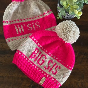 Puede incluir: Dos gorros de punto con pompones. Los gorros son de color rosa y beige con letras blancas. El primer gorro dice "In Style" y el segundo gorro dice "Believe".