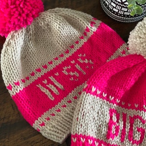 May include: Two knitted hats with pom-poms. The hats are pink and beige with white hearts and the words "Winter" and "Love" knitted into the fabric.