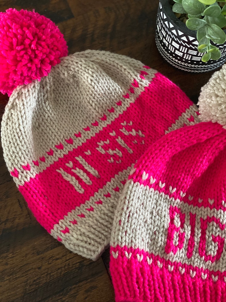 May include: Two knitted hats with pom-poms. The hats are pink and beige with white hearts and the words "Winter" and "Love" knitted into the fabric.