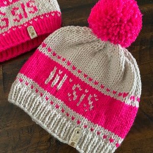 May include: A beige knitted hat with a pink pom pom and a pink and white striped pattern. The hat has the word "Joy" knitted into the pink stripe. The hat has a brown leather tag with the text "Bespoke Knit" on it.