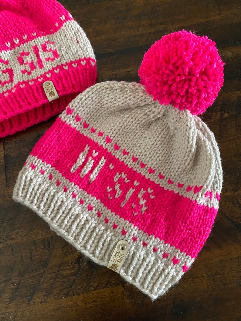 May include: A beige knitted hat with a pink pom pom and a pink and white striped pattern. The hat has the word "Joy" knitted into the pink stripe. The hat has a brown leather tag with the text "Bespoke Knit" on it.