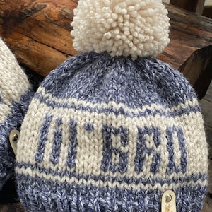 Puede incluir: Gorro de punto azul y blanco con un pomp&oacute;n blanco y la palabra "OUTER" tejida en blanco en la parte delantera. El gorro tiene una etiqueta de cuero con el texto "Brigitte Knit by C. Cuppa".