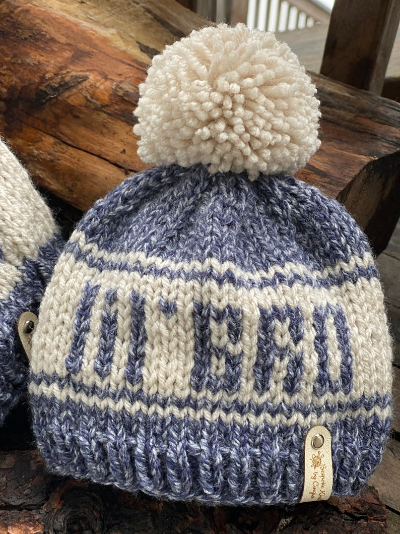 Puede incluir: Gorro de punto azul y blanco con un pomp&oacute;n blanco y la palabra "OUTER" tejida en blanco en la parte delantera. El gorro tiene una etiqueta de cuero con el texto "Brigitte Knit by C. Cuppa".