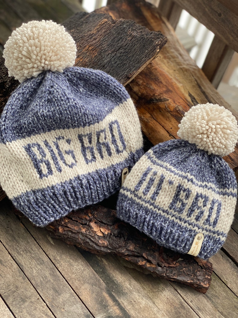 Puede incluir: Dos gorros de punto con pompones. El gorro m&aacute;s grande es azul y blanco con el texto "Big Bro" en la parte delantera. El gorro m&aacute;s peque&ntilde;o es azul y blanco con el texto "Little Bro" en la parte delantera.