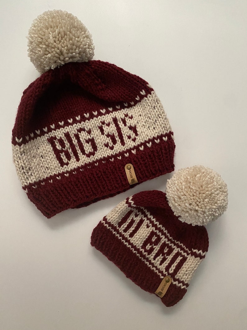 May include: Two knitted beanies with pom poms. The larger beanie is burgundy with white stripes and the text "Big Sis" embroidered on it. The smaller beanie is burgundy with white stripes and the text "Little" embroidered on it.