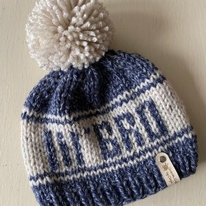 Puede incluir: Gorro de punto azul y blanco con un pomp&oacute;n blanco en la parte superior. El gorro tiene la palabra "Winter" tejida en la banda blanca. Una peque&ntilde;a etiqueta de cuero marr&oacute;n con el texto "Handmade by Cringe" est&aacute; unida al lateral.