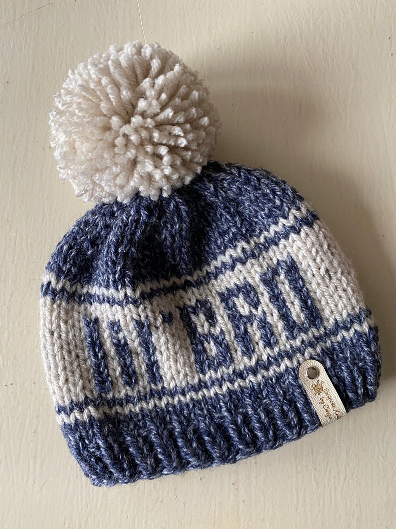 Puede incluir: Gorro de punto azul y blanco con un pomp&oacute;n blanco en la parte superior. El gorro tiene la palabra "Winter" tejida en la banda blanca. Una peque&ntilde;a etiqueta de cuero marr&oacute;n con el texto "Handmade by Cringe" est&aacute; unida al lateral.