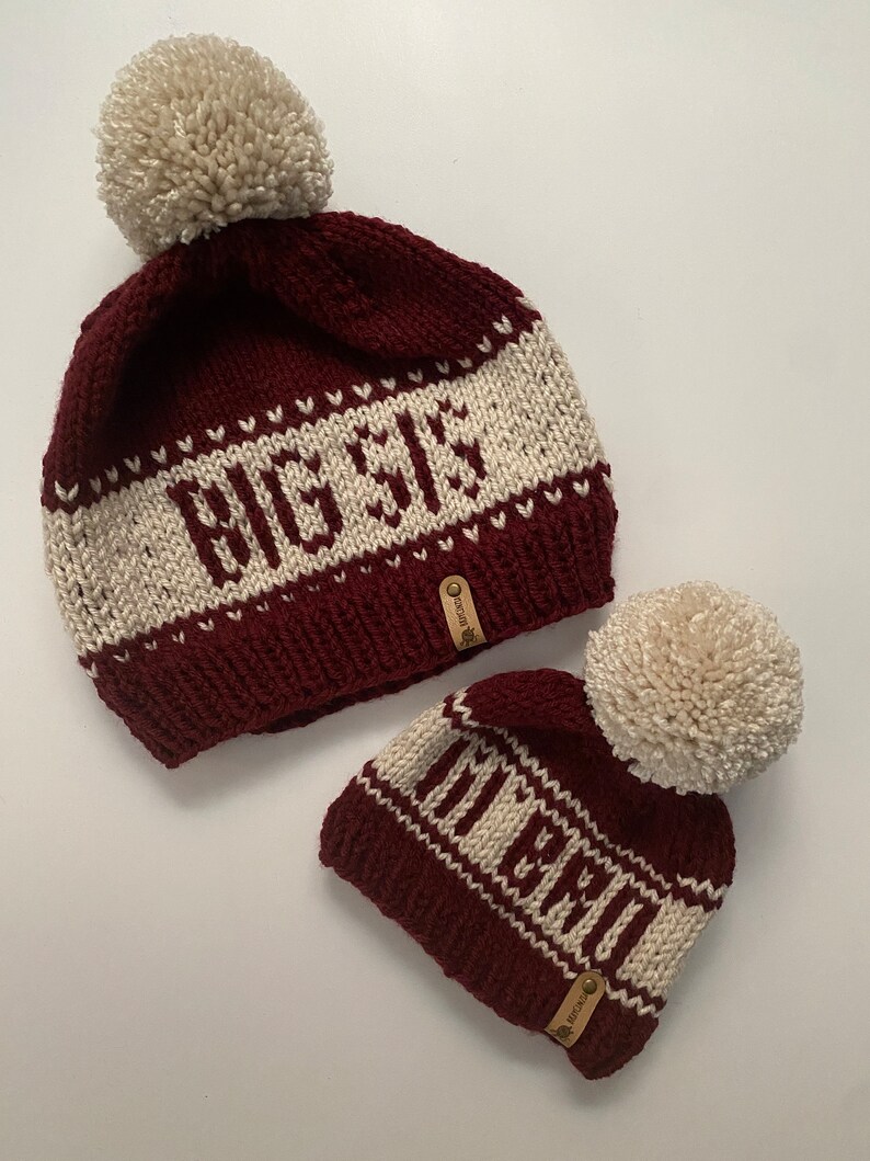 Puede incluir: Dos gorros de punto con pompones. El gorro m&aacute;s grande es burdeos con rayas blancas y el texto "Big Sis" bordado en &eacute;l. El gorro m&aacute;s peque&ntilde;o es burdeos con rayas blancas y el texto "Little" bordado en &eacute;l.
