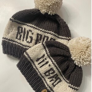 Puede incluir: Dos gorros de punto con pompones. El gorro m&aacute;s grande es gris con rayas blancas y el texto "BIG MOM" en blanco. El gorro m&aacute;s peque&ntilde;o es gris con rayas blancas y el texto "MY BABY" en blanco.
