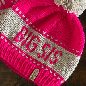 May include: A pink knit beanie with a white pom pom and a white stripe with the word "Lauren" in a white knit font. The beanie has a small brown leather tag that says "Bespoke Knits".