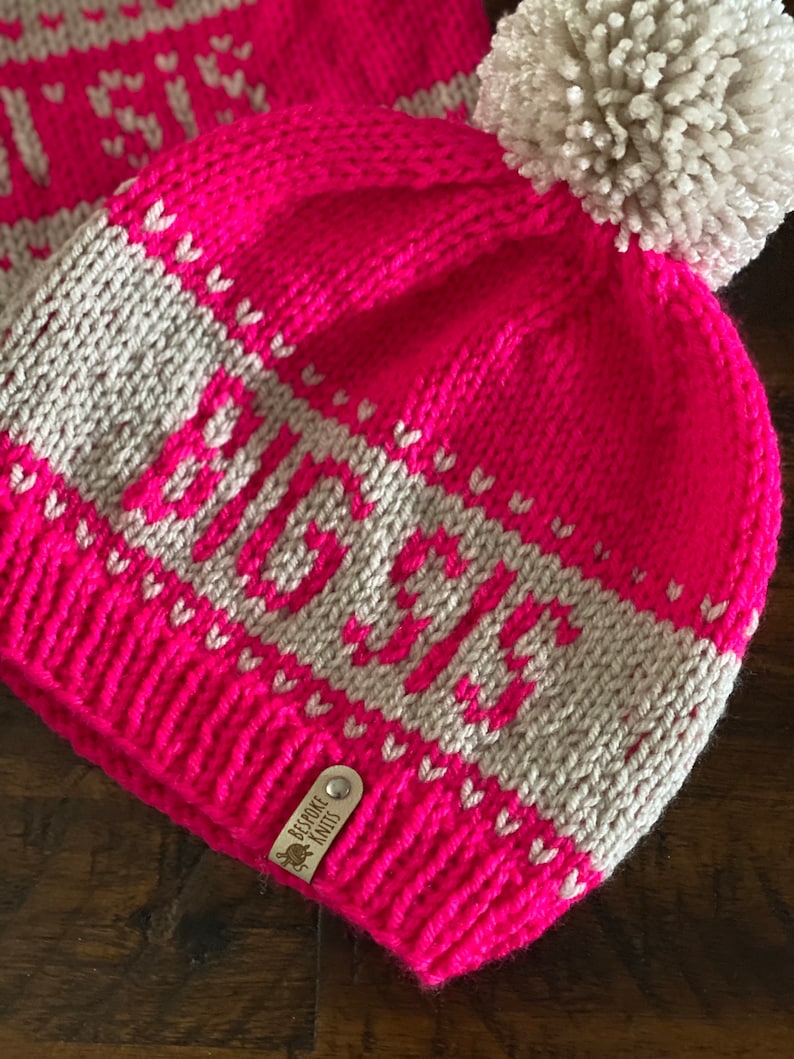 May include: A pink knit beanie with a white pom pom and a white stripe with the word "Lauren" in a white knit font. The beanie has a small brown leather tag that says "Bespoke Knits".