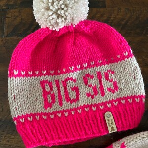 May include: Pink knitted hat with a white pom pom and a white knit band with the word "BEBE" in grey. The hat has a brown leather tag with the text "BESPOKE KNITS".