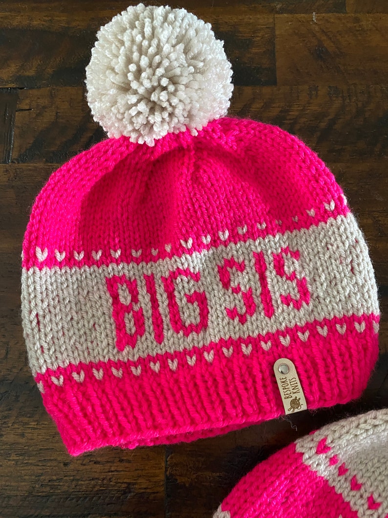 May include: Pink knitted hat with a white pom pom and a white knit band with the word "BEBE" in grey. The hat has a brown leather tag with the text "BESPOKE KNITS".