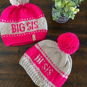 May include: Two knitted hats with pom-poms. One hat is pink with a white pom-pom and says "BIG SIS" in white letters. The other hat is beige with a pink pom-pom and says "MY SIS" in pink letters.