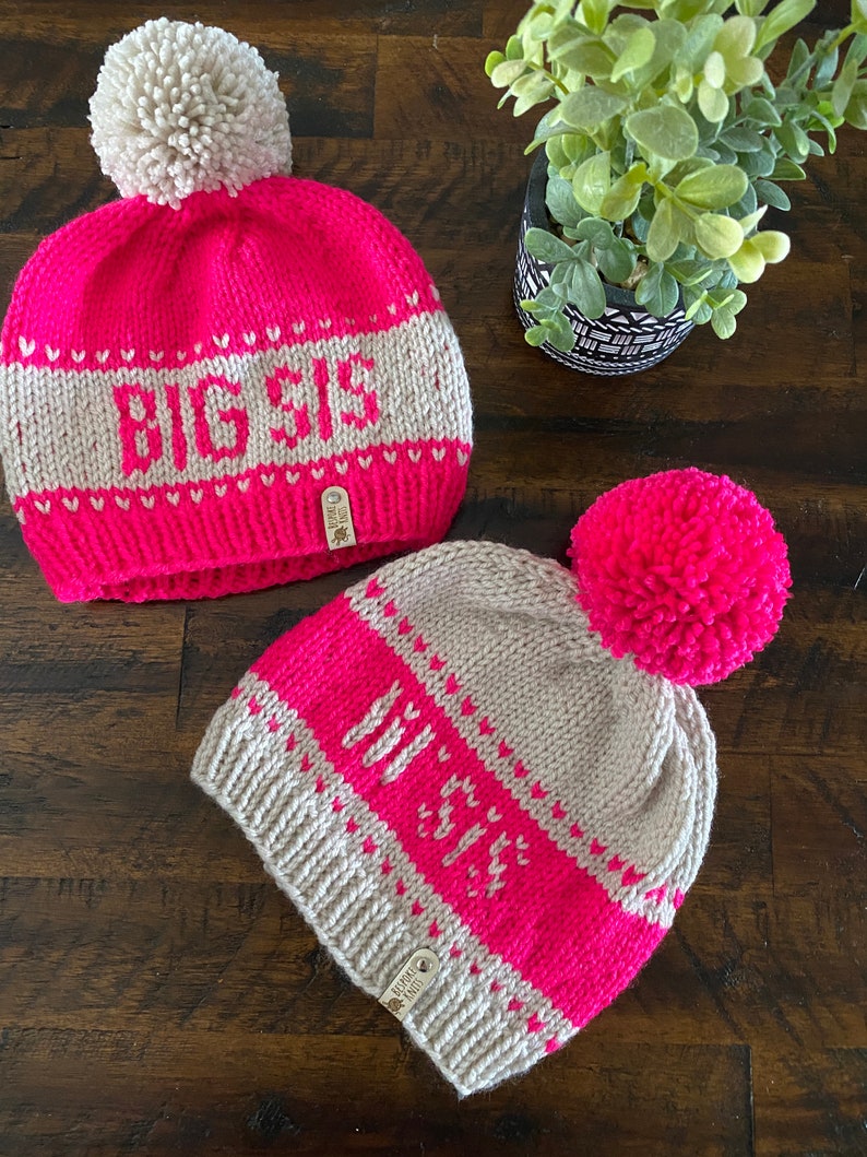 May include: Two knitted hats with pom-poms. One hat is pink with a white pom-pom and says "BIG SIS" in white letters. The other hat is beige with a pink pom-pom and says "MY SIS" in pink letters.