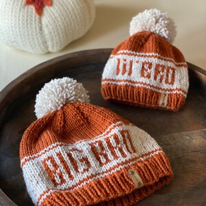 Puede incluir: Dos gorros de punto con pompones. El gorro m&aacute;s grande es marr&oacute;n con rayas blancas y el texto "BEACH" tejido en blanco. El gorro m&aacute;s peque&ntilde;o es marr&oacute;n con rayas blancas y el texto "JU & JU" tejido en blanco.