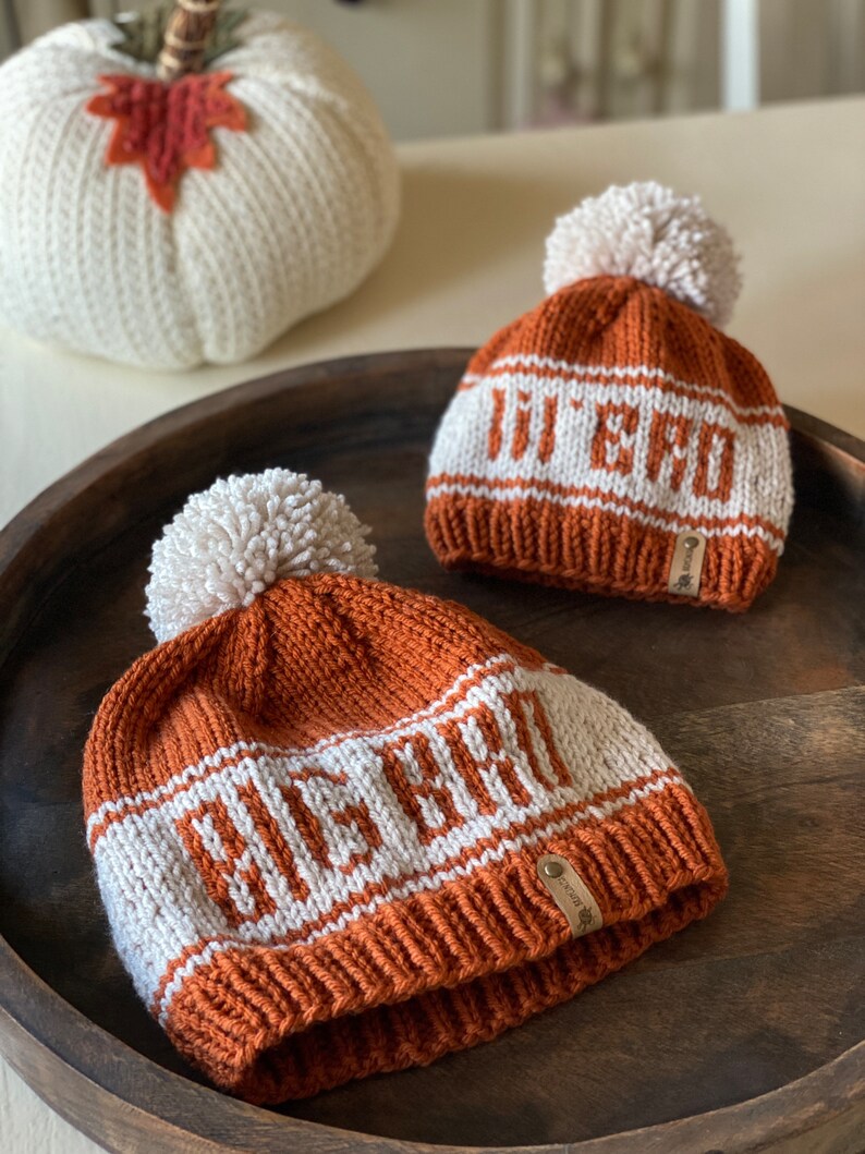 Puede incluir: Dos gorros de punto con pompones. El gorro m&aacute;s grande es marr&oacute;n con rayas blancas y el texto "BEACH" tejido en blanco. El gorro m&aacute;s peque&ntilde;o es marr&oacute;n con rayas blancas y el texto "JU & JU" tejido en blanco.