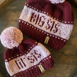 May include: Two knitted hats with pom-poms. The hats are burgundy with a pink stripe and the words "ELISE" and "OLIVIA" knitted in pink.