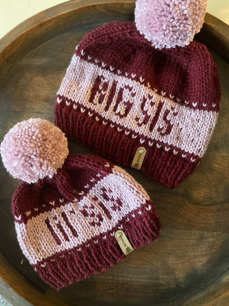 May include: Two knitted hats with pom-poms. The hats are burgundy with a pink stripe and the words "ELISE" and "OLIVIA" knitted in pink.