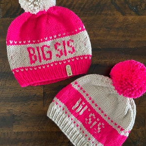 May include: Two knitted hats with pom-poms. The larger hat is pink with a white pom-pom and says "BIG SIS" in white letters. The smaller hat is beige with a pink pom-pom and says "LIT SIS" in white letters.