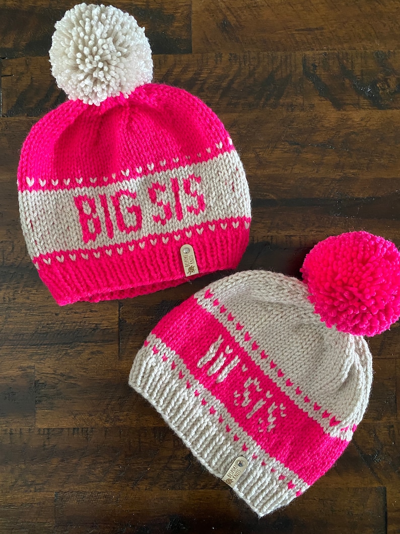 May include: Two knitted hats with pom-poms. The larger hat is pink with a white pom-pom and says "BIG SIS" in white letters. The smaller hat is beige with a pink pom-pom and says "LIT SIS" in white letters.