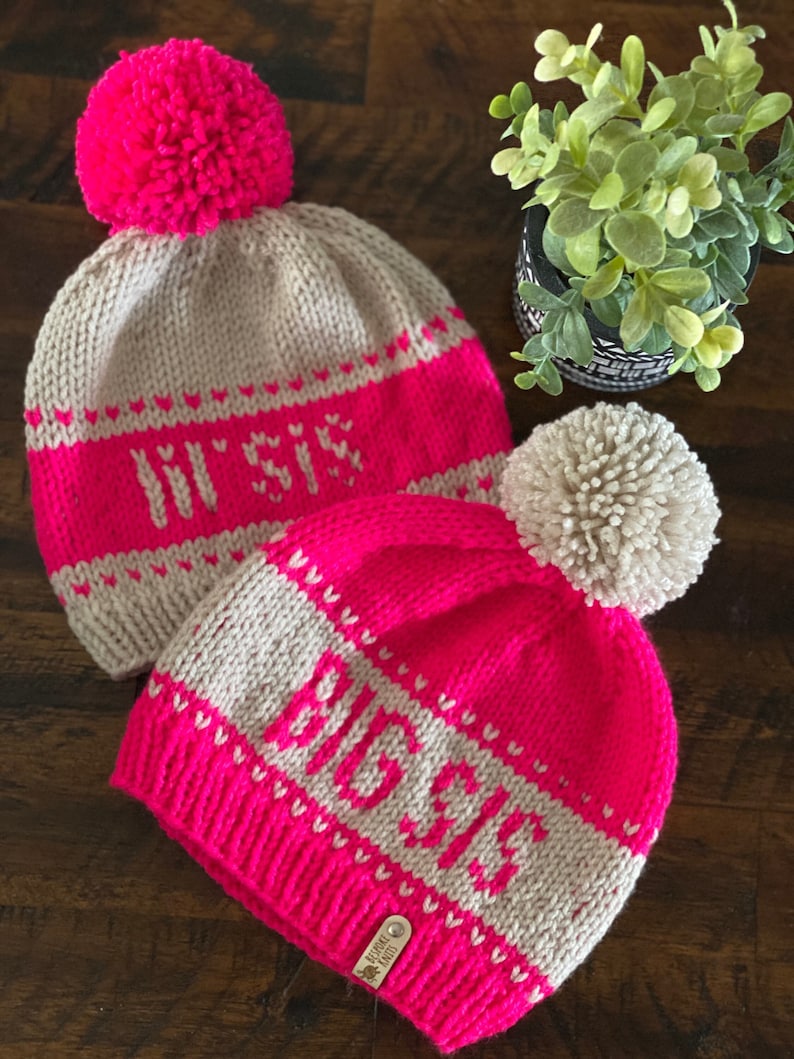 May include: Two knitted hats with pom-poms. The hats are pink and beige with white lettering. The first hat says "In Style" and the second hat says "Believe".