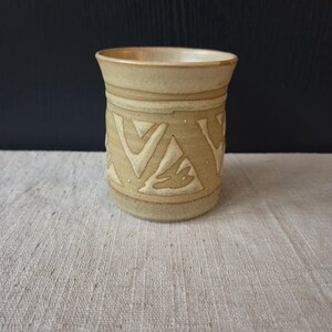 May include: A beige ceramic tumbler with a textured surface and a geometric pattern carved into the exterior. The cup has a slightly flared rim and is set against a neutral background.