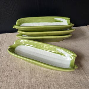 May include: A collection of ceramic corn-shaped serving dishes. The dishes are painted in shades of green and white, with a detailed design mimicking corn on the cob. The dishes are stacked on a neutral-coloured surface.