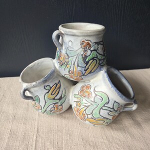 May include: A trio of ceramic mugs with handles, arranged together. The mugs are pale blue with hand-painted floral and figure designs in orange, green, and brown. The mugs have a rustic, handmade appearance.