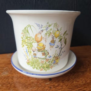 May include: A white ceramic plant pot with a matching saucer. The pot has a whimsical illustration of mice with balloons and floral elements in green, yellow, and blue. The saucer has a blue rim.