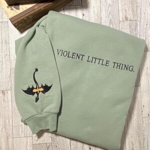 Puede incluir: Una sudadera de color verde salvia con un drag&oacute;n bordado en negro en la manga y el texto "VIOLENT LITTLE THING." en la parte delantera.