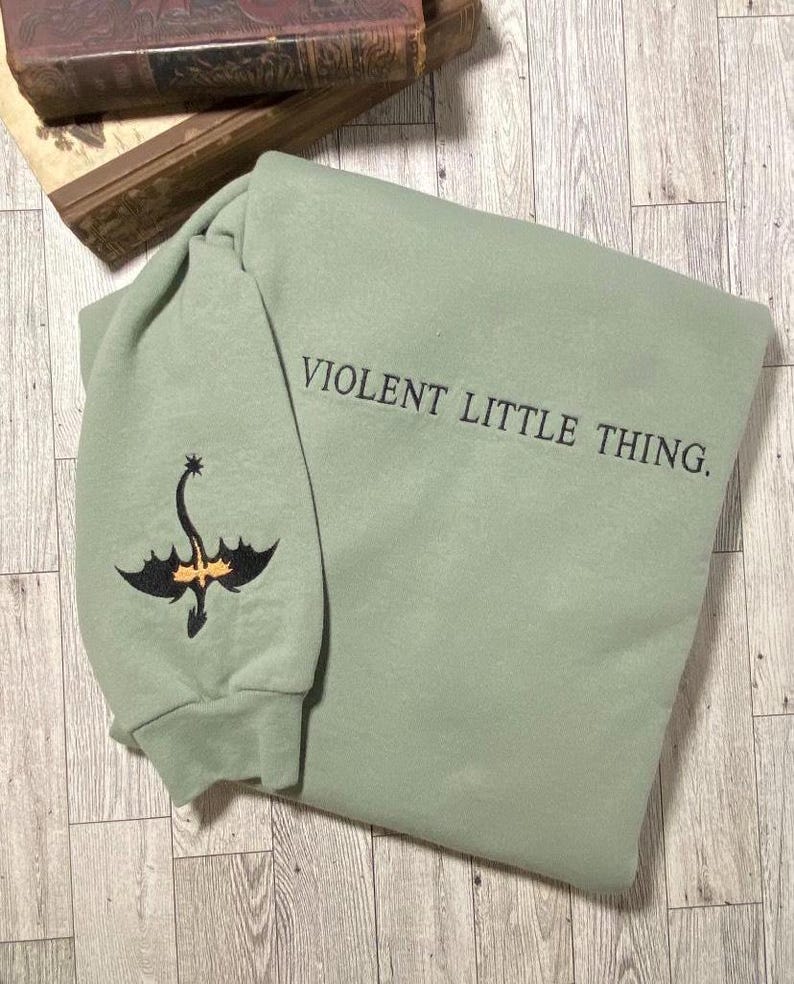 Puede incluir: Una sudadera de color verde salvia con un drag&oacute;n bordado en negro en la manga y el texto "VIOLENT LITTLE THING." en la parte delantera.