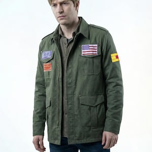 Puede incluir: Chaqueta verde oliva de estilo militar con múltiples bolsillos y parches, incluida una bandera estadounidense. La chaqueta tiene una parte delantera abotonada y un cuello. La persona lleva debajo una camisa marrón abotonada.
