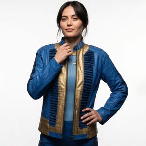 May include: A blue and gold jacket featuring a high collar, vertical pleated dark blue panels along the front, and a central gold zipper closure. The garment is constructed with smooth blue panels on the sleeves and torso, accented by metallic gold trim.