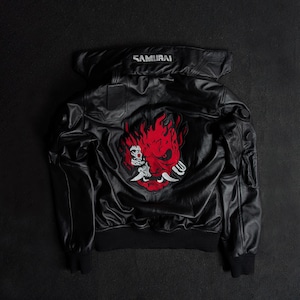 May include: A black leather jacket featuring a large red and white graphic of a stylized robotic demon face on the back. The collar area displays the word SAMURAI in white text. The jacket includes ribbed cuffs and hem, with structured paneling.