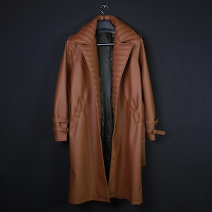 May include: A long, tan-colored trench coat featuring a wide, quilted lapel design, long sleeves with adjustable buckled straps at the cuffs, and a dark interior lining. The coat is displayed hanging on a hanger against a plain dark background.
