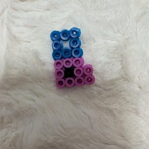 Op de afbeelding: Een kleine, handgemaakte letter "L" gemaakt van blauwe en paarse kralen. Het bovenste gedeelte is blauw, het onderste gedeelte is paars en het midden is zwart. De kralenletter wordt weergegeven op een zacht, wit oppervlak.